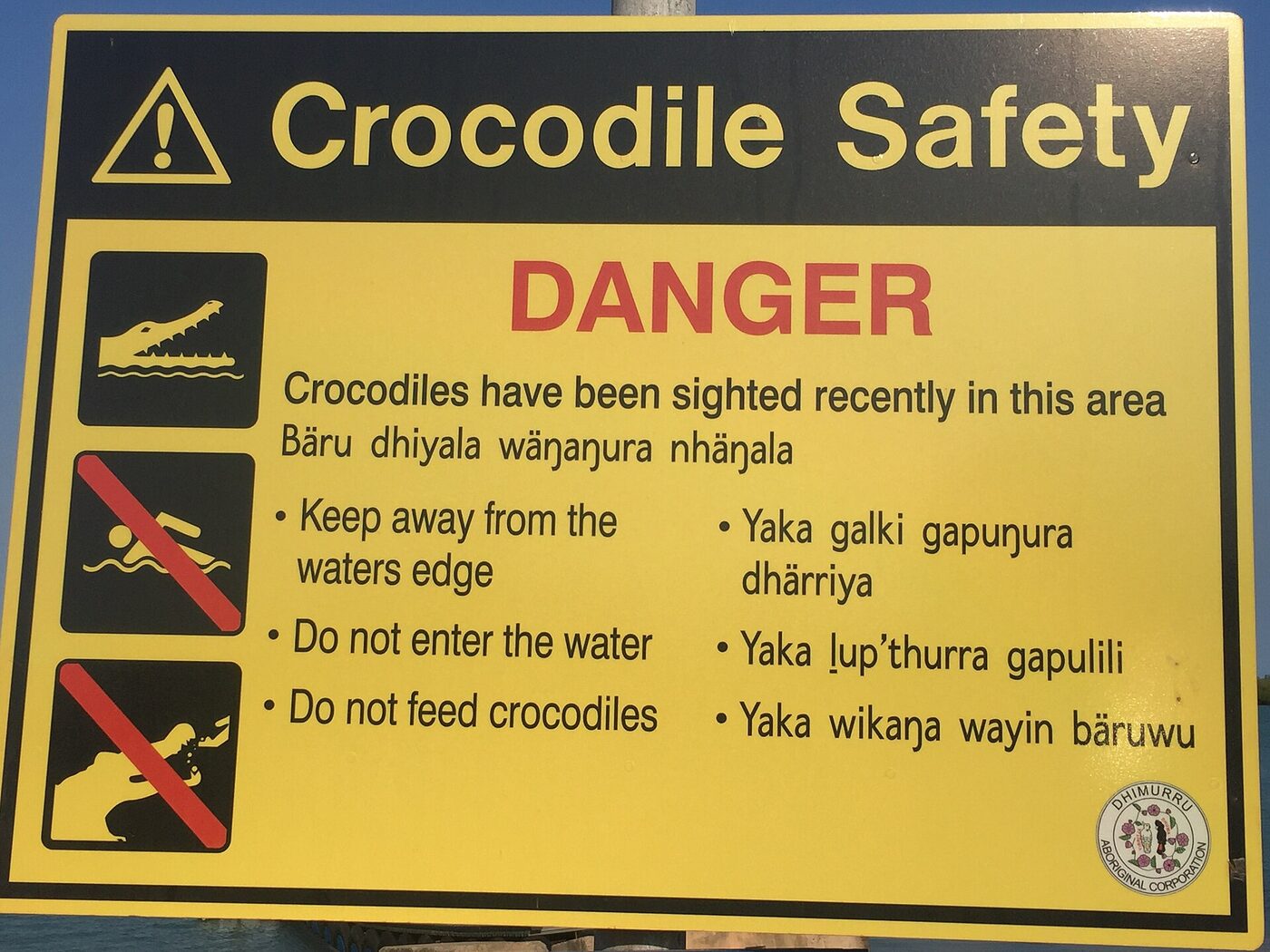 A yellow and black crocodile safety sign headed DANGER, with parallel warnings in English and Yolŋu Matha; the Yolŋu lines contain the letter eng, vowels with a diaeresis and an underlined letter l.