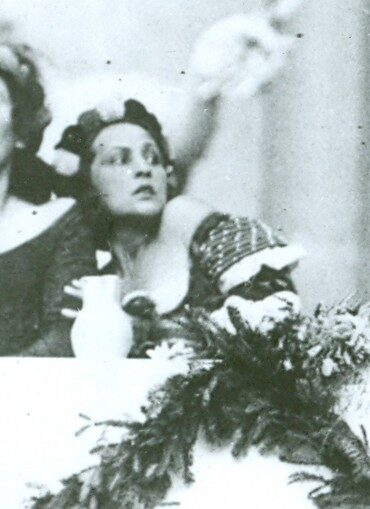 A grainy black-and-white photograph cropped close on a costumed woman leaning back from a white-clothed table set with a jug and a spray of greenery, another performer's raised arm behind her.