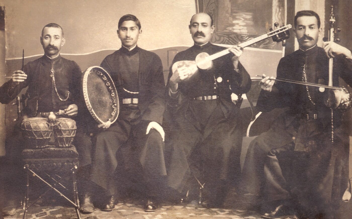 Sepia studio photograph of four seated musicians in long dark coats, holding, from the left, a pair of small drums with sticks, a large frame drum, a long-necked plucked lute and a bowed spike fiddle.