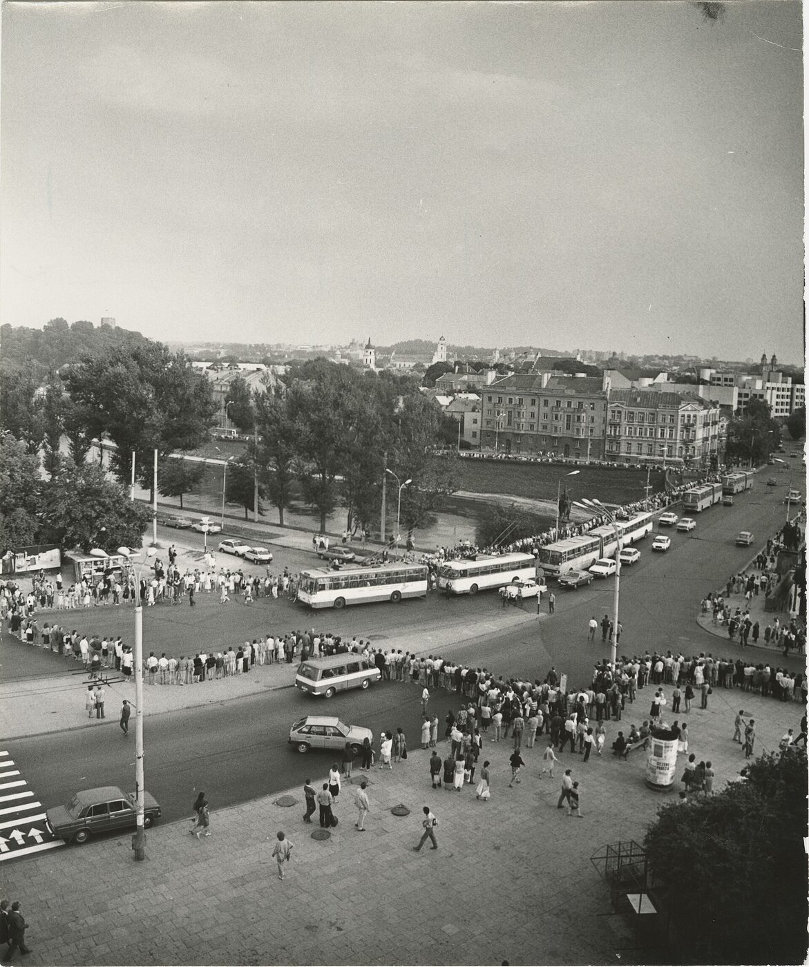 A black-and-white photograph looking down on a city crossroads: a single continuous line of people stands along the pavements and across the junction, past halted buses and trolleybuses and parked cars, with a river embankment, trees, apartment blocks and church towers behind.