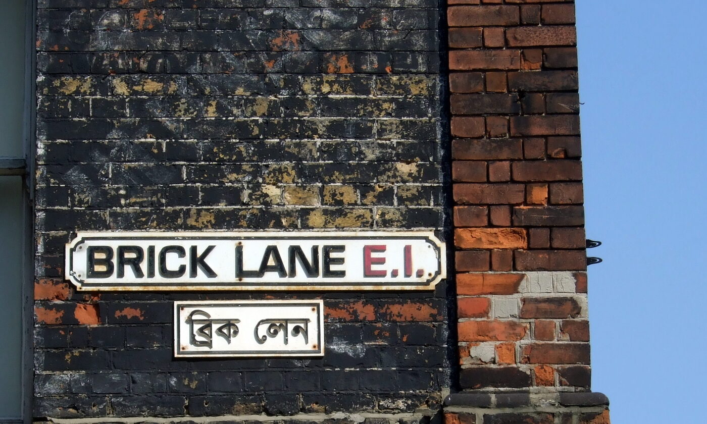 Two street nameplates on a brick wall: the upper reads Brick Lane in roman capitals, the lower repeats the name in Bengali script.
