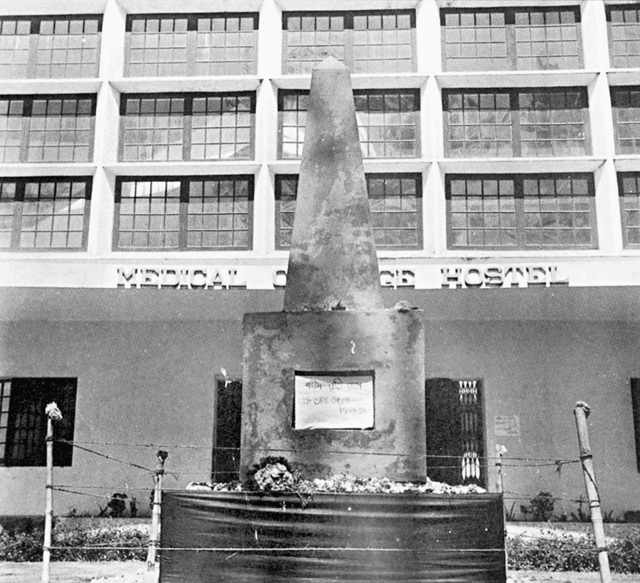 A tall narrow monument on a plinth, hung with a placard in Bengali and banked with flowers, standing before a multi-storey college building in a grainy black-and-white photograph.