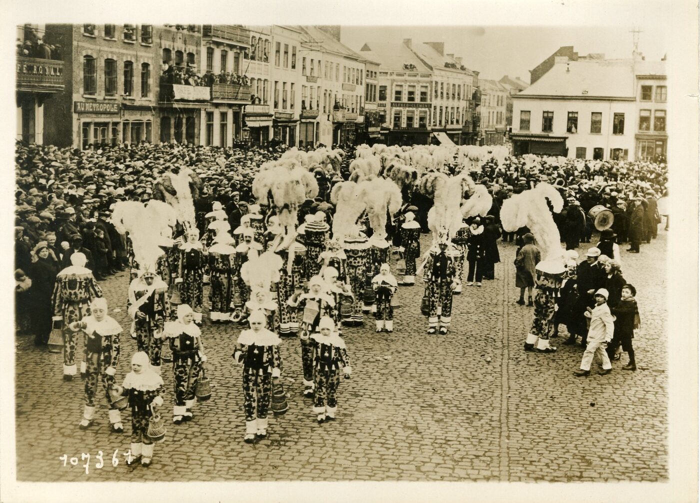 Black and white photograph of a cobbled market square packed with spectators, through which walk dozens of costumed figures in patterned suits and white ruffs, many wearing tall plumed ostrich-feather headdresses and carrying baskets.