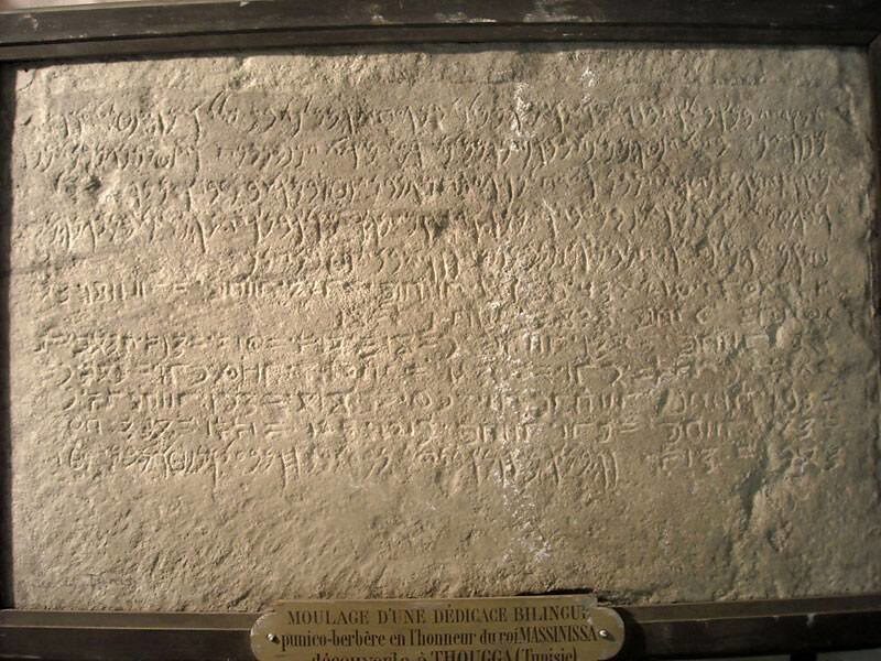 A framed plaster cast of a stone slab densely incised with several lines of two different ancient scripts, with a museum label fixed below it.