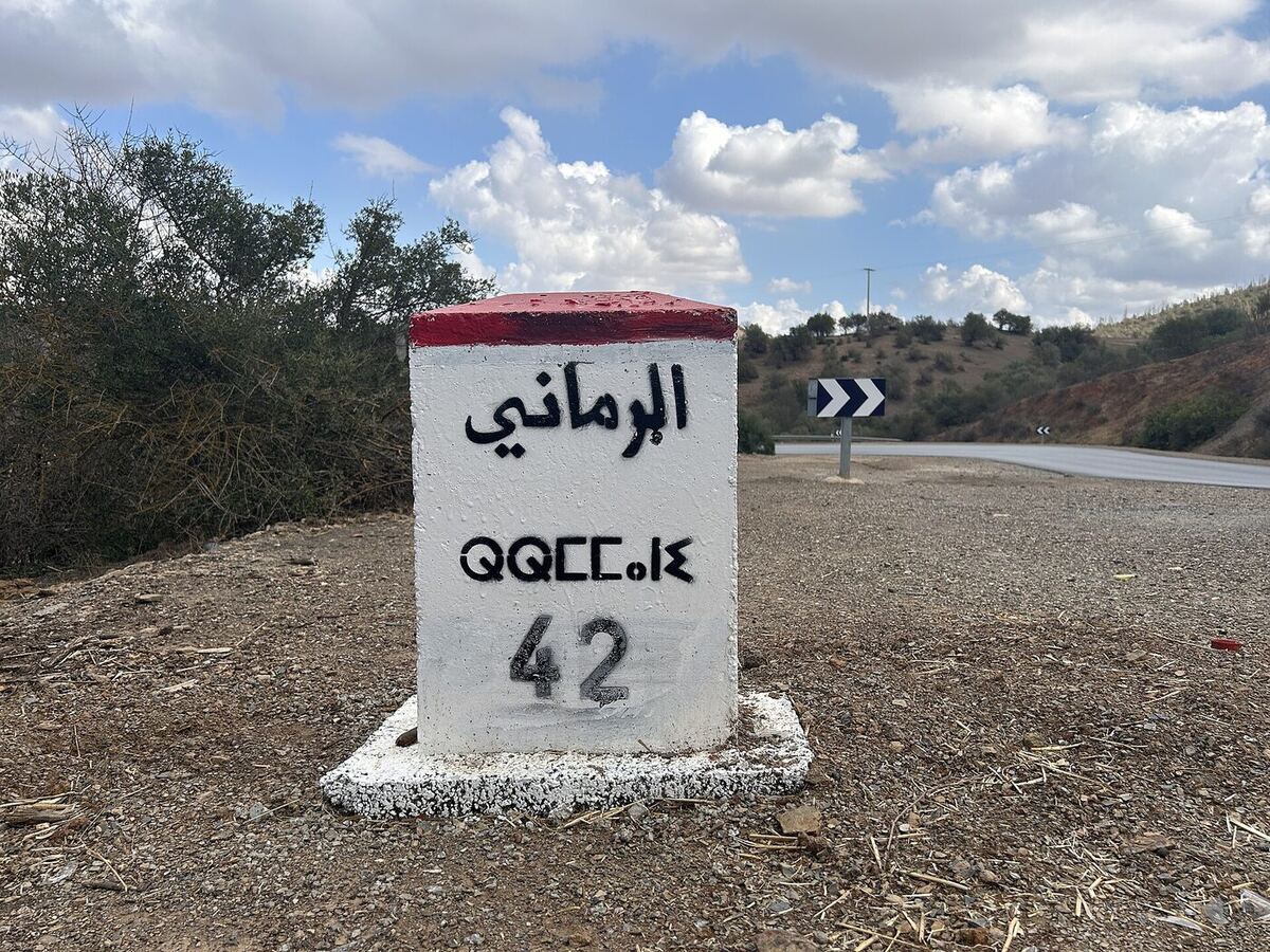 A whitewashed red-topped roadside kilometre stone beside a rural road, painted with a place name in Arabic script, the same name in Tifinagh characters below it, and the number 42.