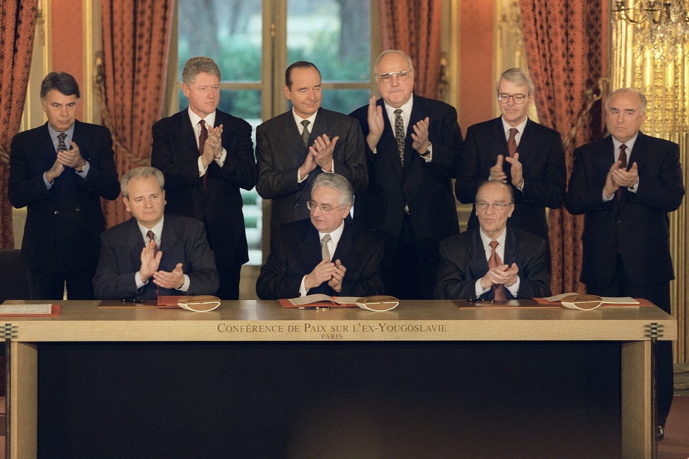 Nine men in dark suits in a gilded room, three seated at a long table with open documents before them and six standing behind; all nine are applauding. The front of the table is inscribed Conference de Paix sur l'ex-Yougoslavie, Paris.