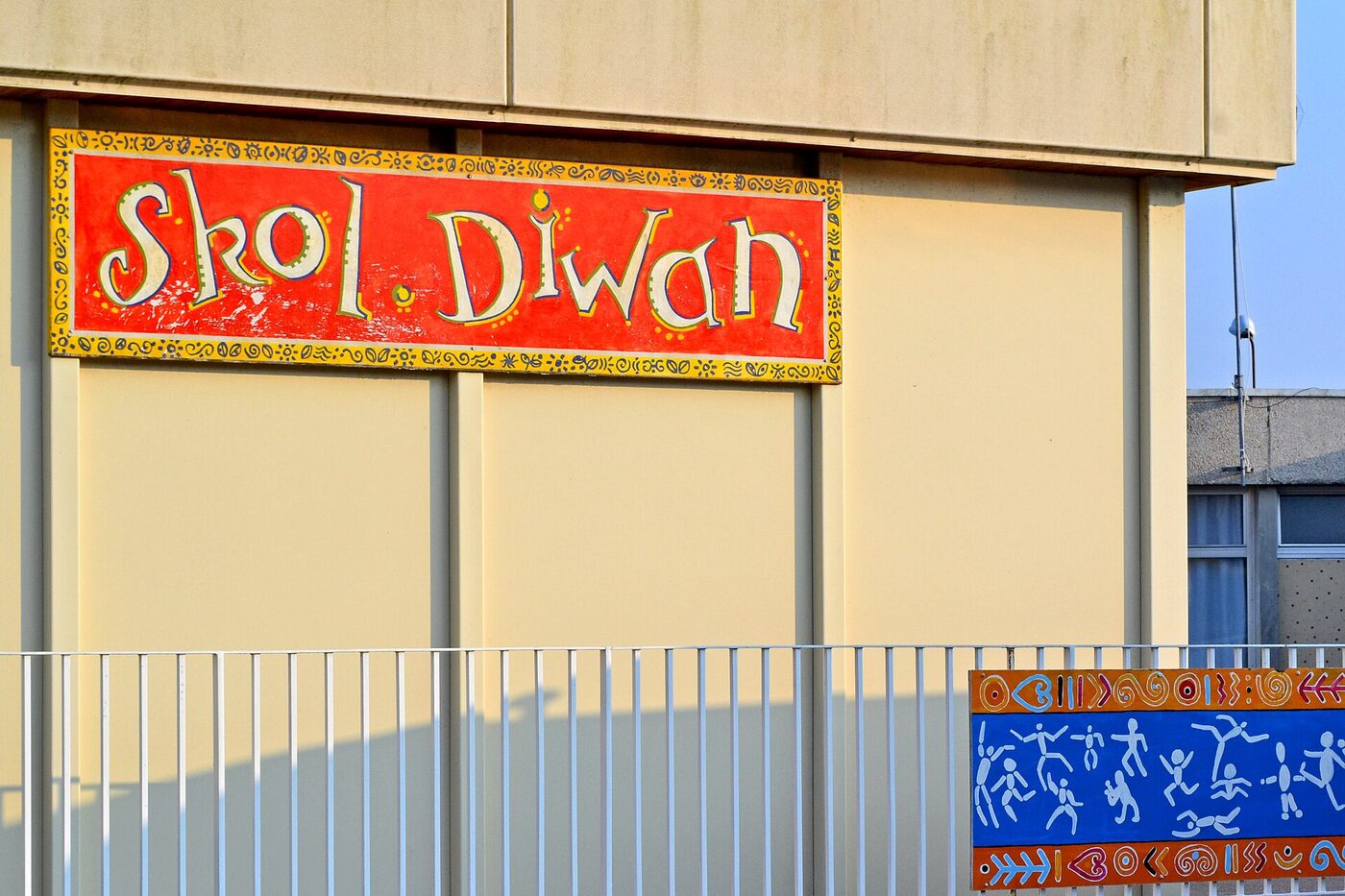 A hand-painted red and yellow sign reading Skol Diwan, in decorated lettering, fixed to the wall of a modern school building.