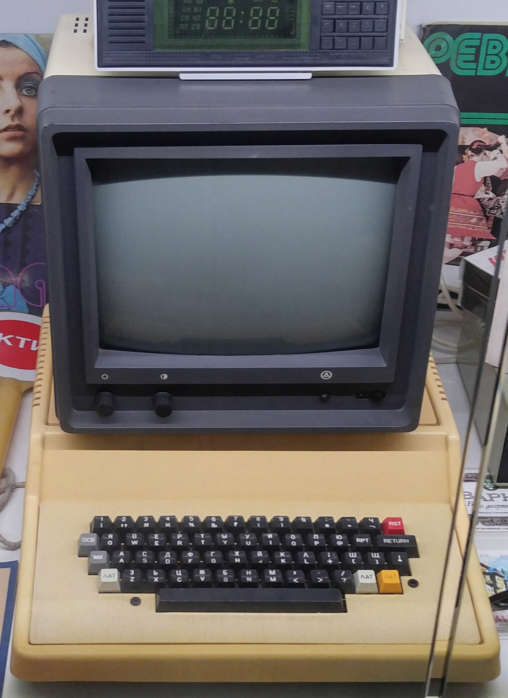 A beige wedge-shaped microcomputer with an integrated keyboard and a grey monitor sitting on top of it, displayed in a museum. Each keycap carries a Cyrillic letter above a Latin one.