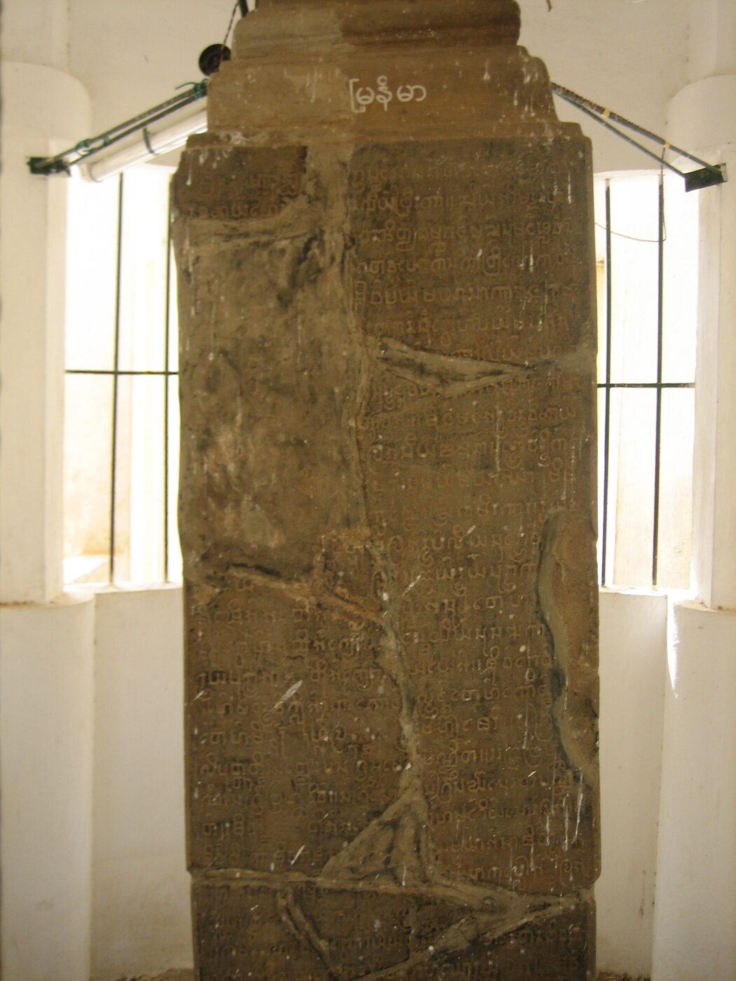 A cracked and mended stone pillar densely incised with rows of rounded Burmese characters, standing between barred windows inside a whitewashed shelter.