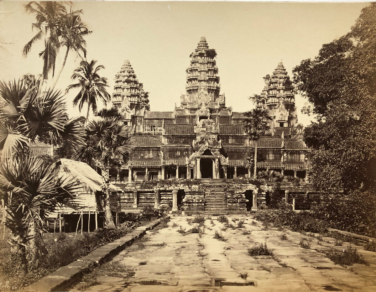 Sepia photograph of Angkor Wat seen along a broken, weed-grown stone causeway, three towers rising behind the terraced galleries, with palms and a thatched hut on stilts at the left.
