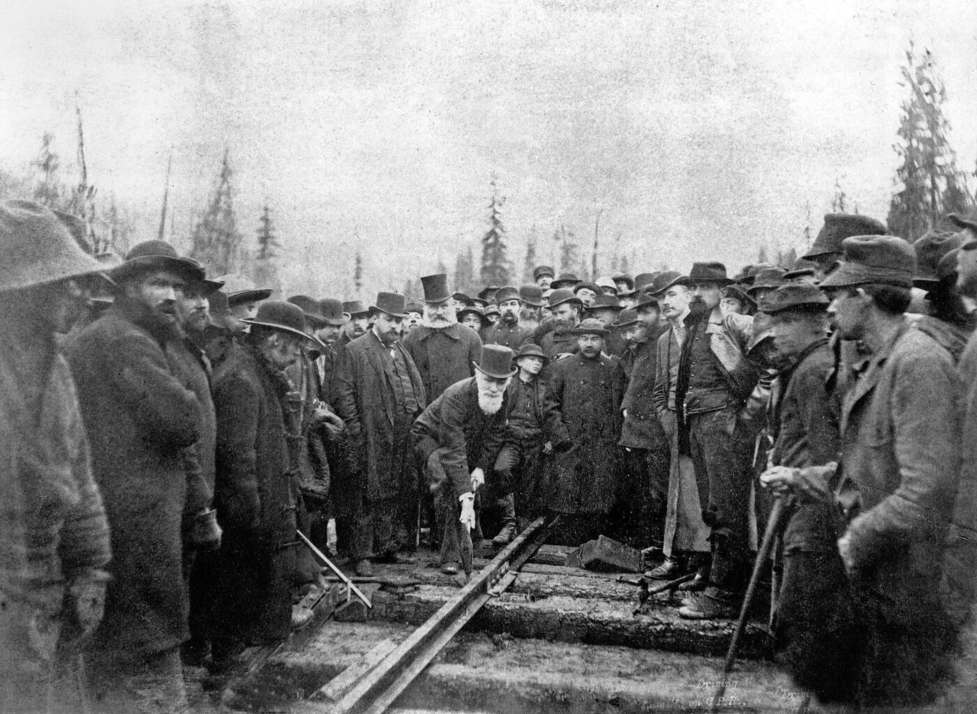 Sepia photograph of a crowd of nineteenth-century railway workers and officials packed around a track in a forest cutting; a bearded man in a long coat and top hat stoops to drive a spike into the rail while a boy in a light cap watches from behind.