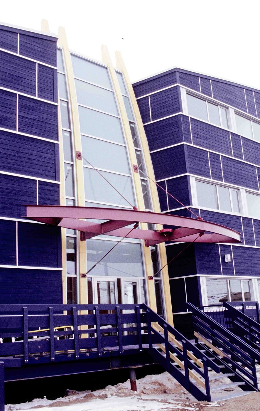 A dark blue clapboard-clad building on piles above snow, its glazed entrance bay framed by two tall pale laminated timber ribs that curve upward past the roofline, with a dark red cantilevered canopy on tie rods over the doors and a blue steel stair and ramp in front.