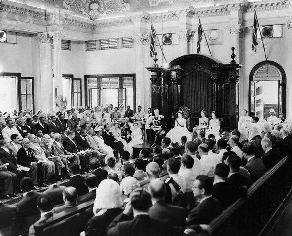 Black and white photograph of a packed colonial-era legislative chamber, flags standing behind a dais, a small ceremonial party seated at the front facing rows of formally dressed guests.
