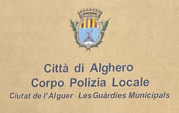 A painted wall sign showing a coat of arms above two lines of Italian text, with the same wording repeated below in Catalan in smaller italic lettering.