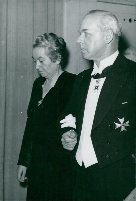 Black-and-white press photograph of a woman in a dark dress walking arm in arm with a man in white tie wearing a breast star and a neck decoration, in front of a draped curtain.