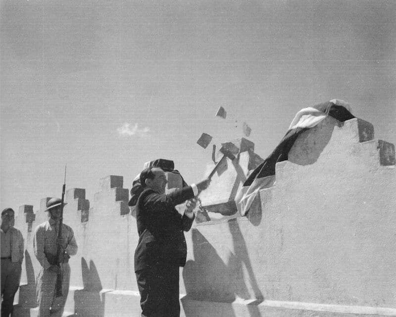 Black and white photograph of a man in a dark suit swinging a sledgehammer at the crenellated top of a whitewashed fortress wall, masonry fragments flying, a draped flag over the wall beside him, and a helmeted soldier with a rifle and another man standing at the foot of the wall.