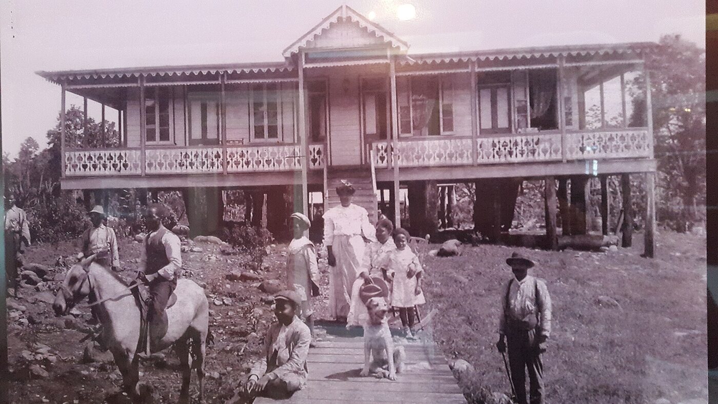 Sepia photograph of a family before a large timber house raised on stilts with a wide verandah and fretwork railings; a woman in a long white dress and hat stands with children in front of the steps, a man sits on a horse at the left with another standing at its head, a third man stands at the right, and a boy and a dog are on a plank walkway.