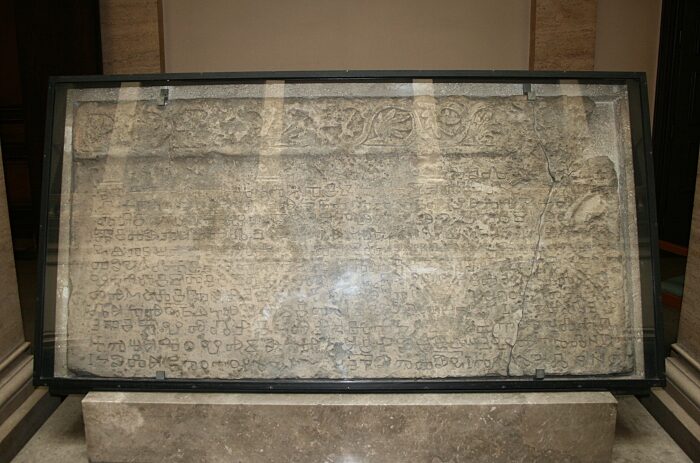 A wide, worn limestone slab inside a glass display case on a stone plinth in a hall, its face covered with rows of angular incised Glagolitic letters under a carved vine border.