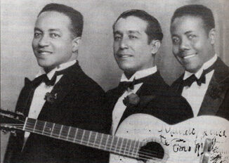 Sepia studio portrait from about 1930 of the three members of the Trio Matamoros in dinner jackets and bow ties, the neck of a guitar visible at the left and a signature inked across the lower corner.