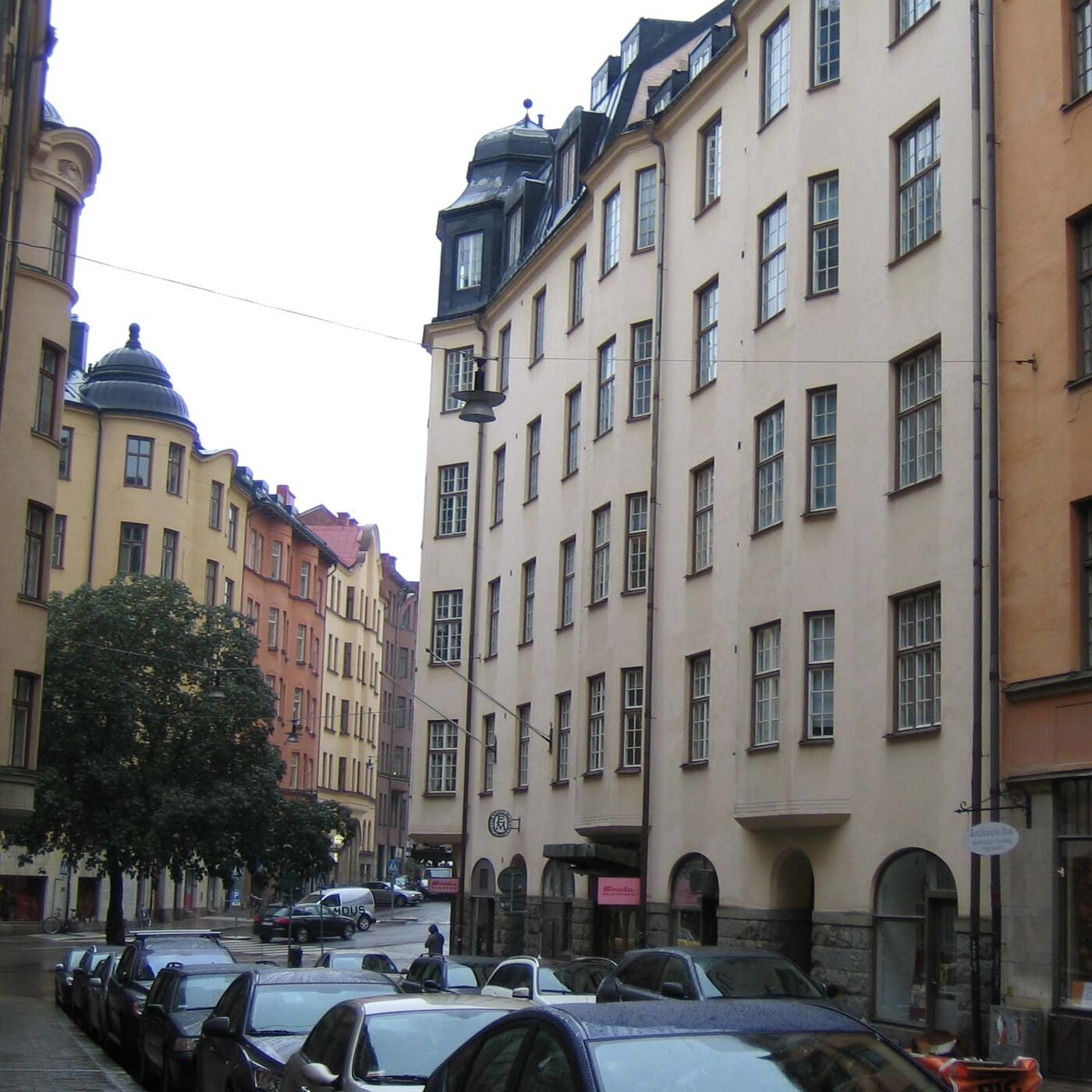 A tall pale rendered building running along a Stockholm street, with arched shopfronts at pavement level, dormer windows in the roof and cars parked along the kerb.