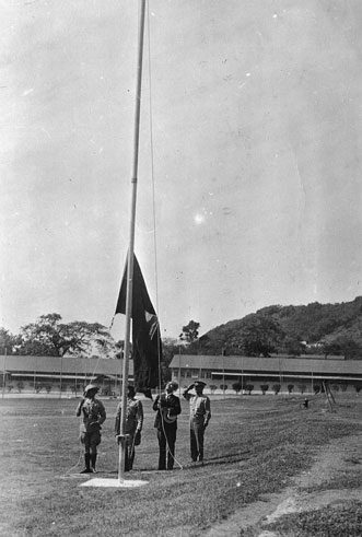 A black and white photograph of a parade ground: four uniformed men stand at the foot of a tall flagpole, one of them saluting, as a flag rises part way up the halyard, with long low barrack buildings and wooded hills behind.