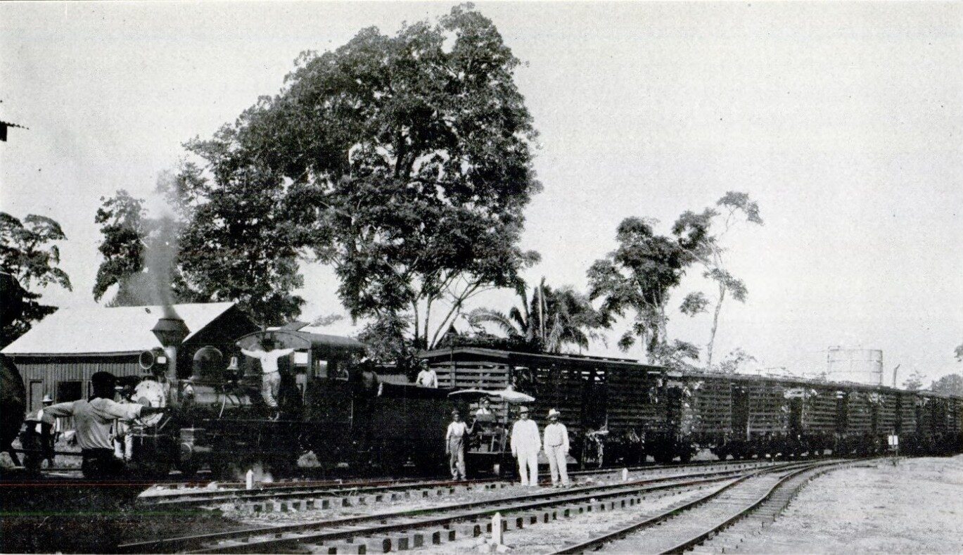Black and white photograph of a small steam locomotive and a line of open slatted freight wagons stopped beside a low wooden station building, with tall trees behind and several men standing on the track.