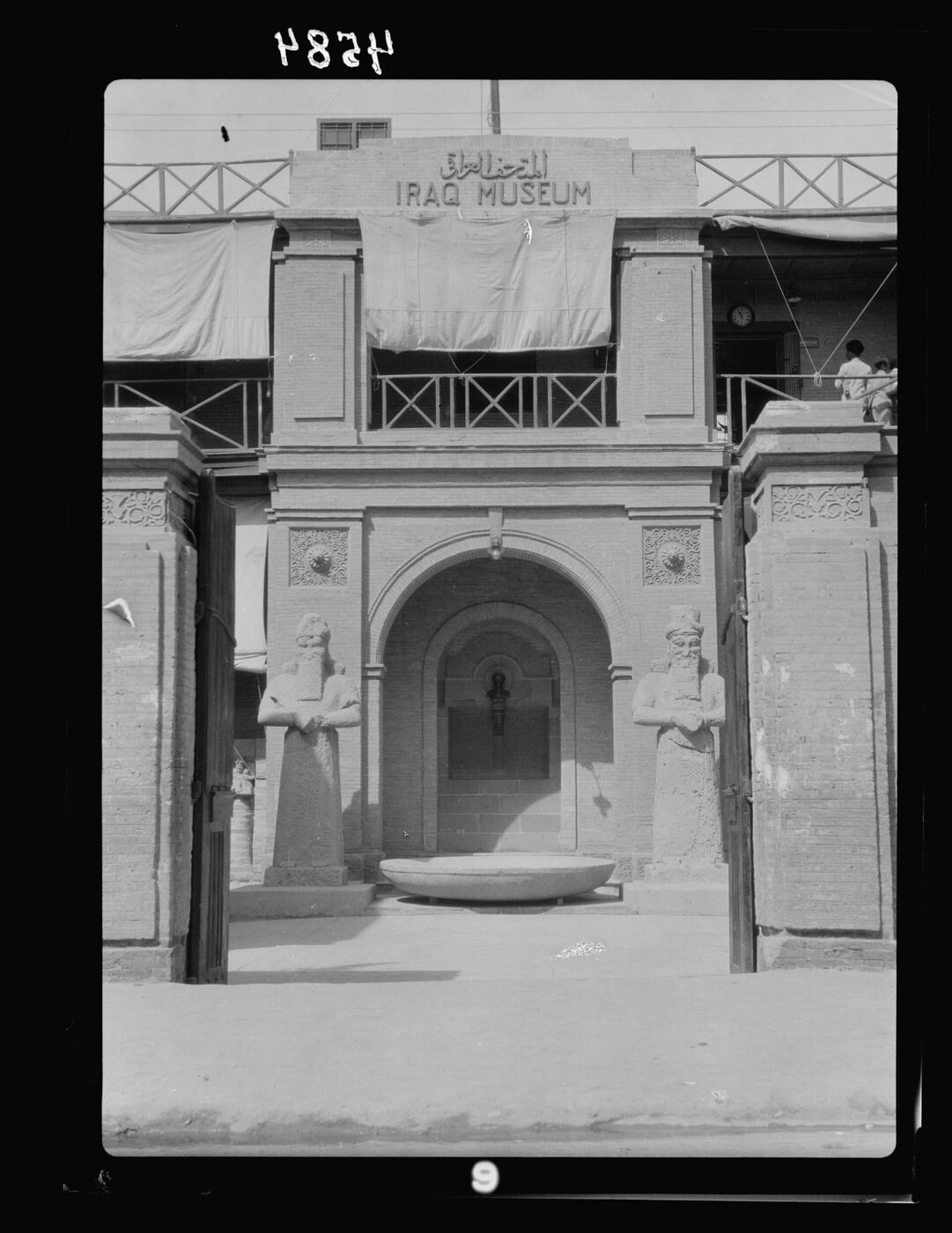 Black-and-white photograph of a gateway lettered IRAQ MUSEUM, with two ancient stone statues standing either side of the arched doorway.
