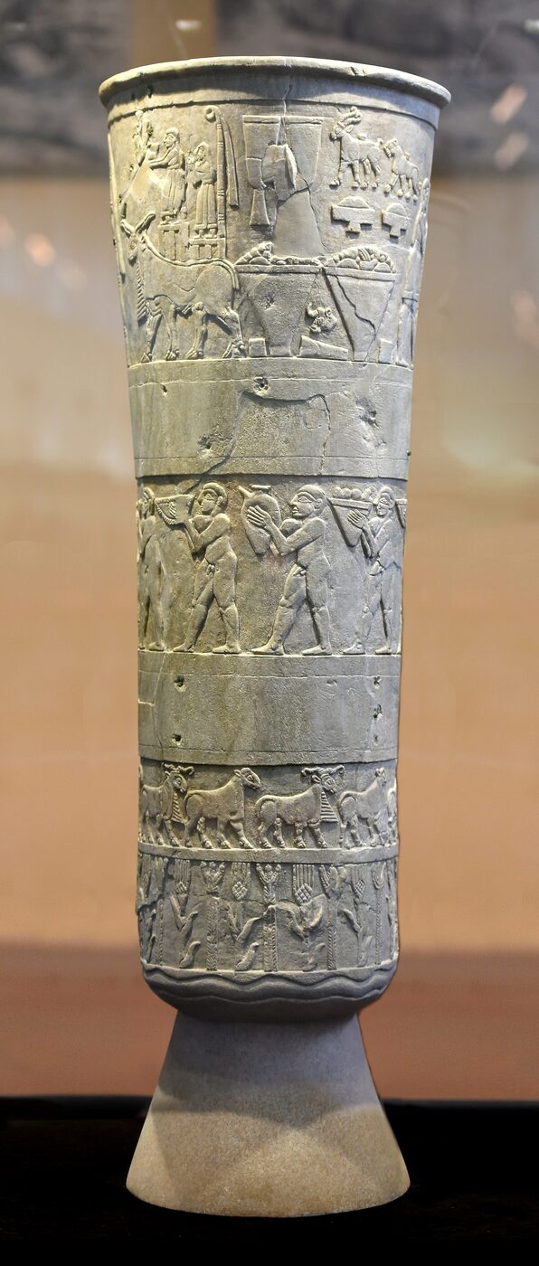 A tall carved alabaster vase with horizontal bands of relief showing plants, animals and human figures, photographed against a plain background.