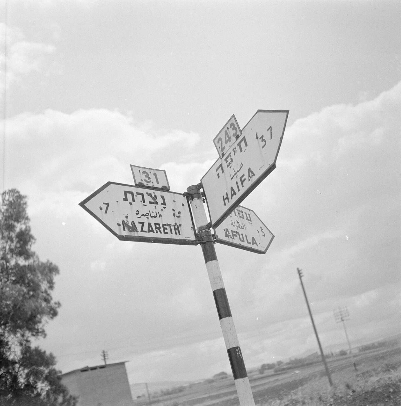 A black and white photograph of a road signpost with four arrow-shaped plates, each carrying a place name in Hebrew with vowel points, in Arabic, and in capital Latin letters reading NAZARETH, HAIFA and AFULA.