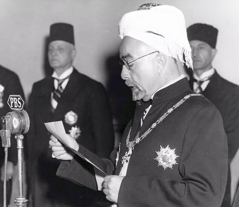A bearded man in glasses, ceremonial robes and a decorated sash reads from a sheet of paper into a large studio microphone, with officials in tarbushes standing behind him.