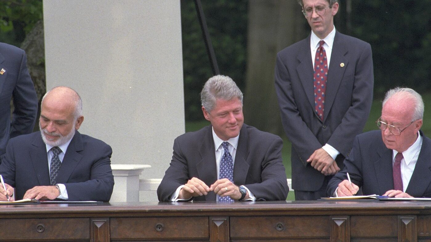 Three men at an outdoor table on a lawn, two seated and signing documents while a third stands between them watching.