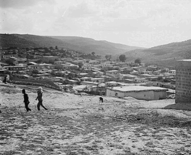 A monochrome photograph looking down a bare slope towards a village of low flat-roofed houses spread across a hillside, two figures walking in the foreground and open hills behind.