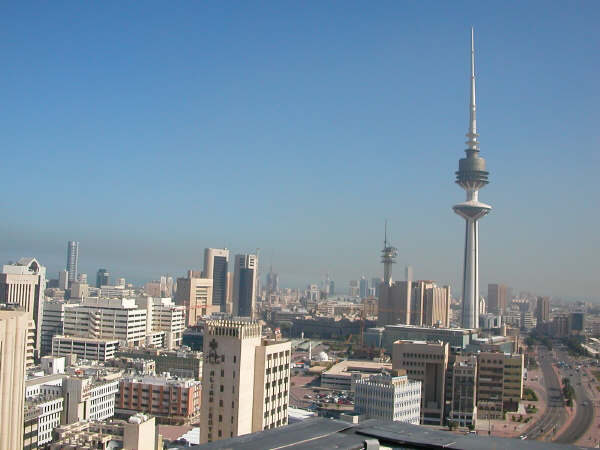 A tall slender telecommunications tower with a banded shaft and disc-shaped observation levels standing above the low white buildings of central Kuwait City under a clear sky.
