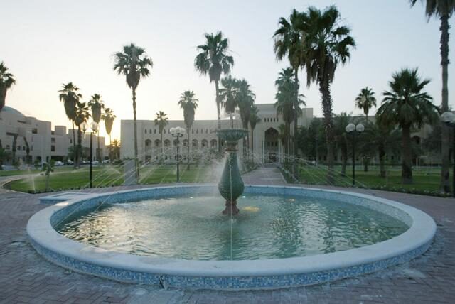 A circular stone fountain playing in a lawned quadrangle, with sprinklers, date palms and low arcaded university buildings behind it in evening light.