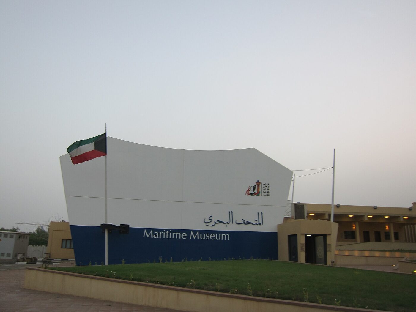 A museum building beside a lawn, its white and blue front curved like a ship's hull and lettered Maritime Museum in Arabic and English, with the Kuwaiti flag flying beside it.