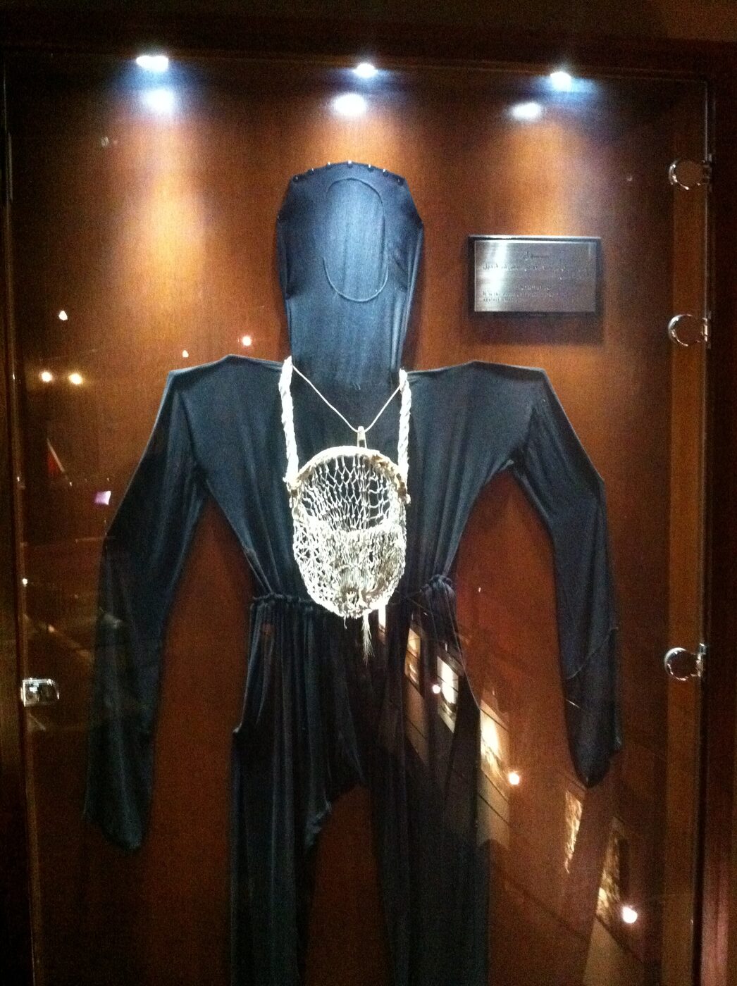 A dark long-sleeved one-piece diving garment with a hood, displayed upright in a glass case with a knotted net bag hanging at its chest.