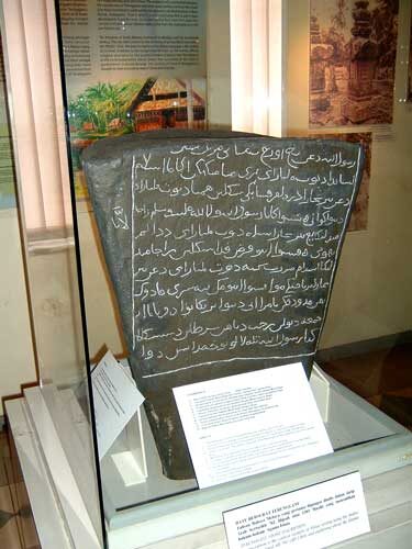 An upright stone slab with several lines of Arabic-script writing carved into its face, standing on a plinth in a museum gallery.