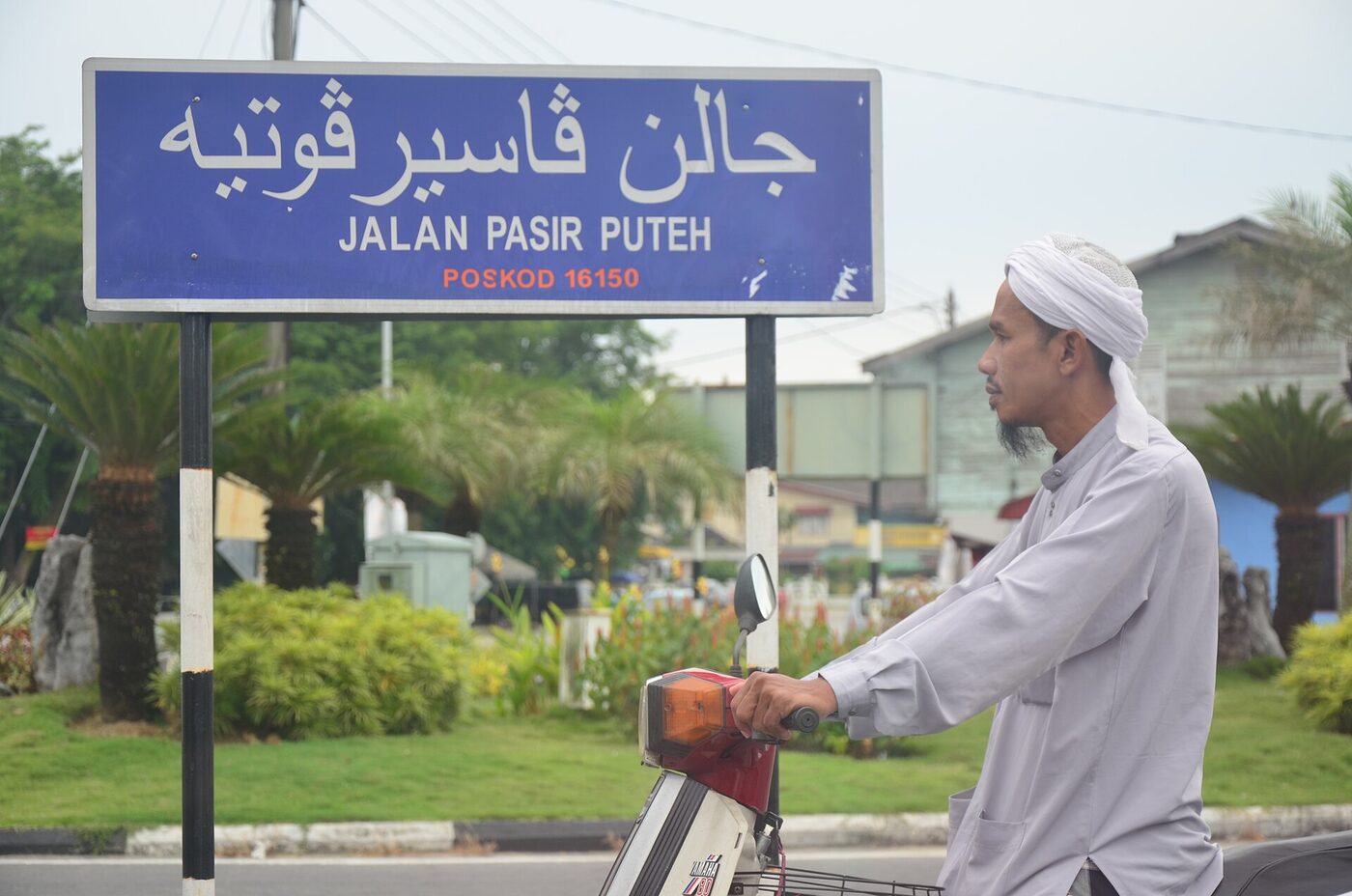 A blue street-name sign carrying the same road name twice, once in Arabic-script Jawi above and once in Latin letters below, with a man standing beside it.