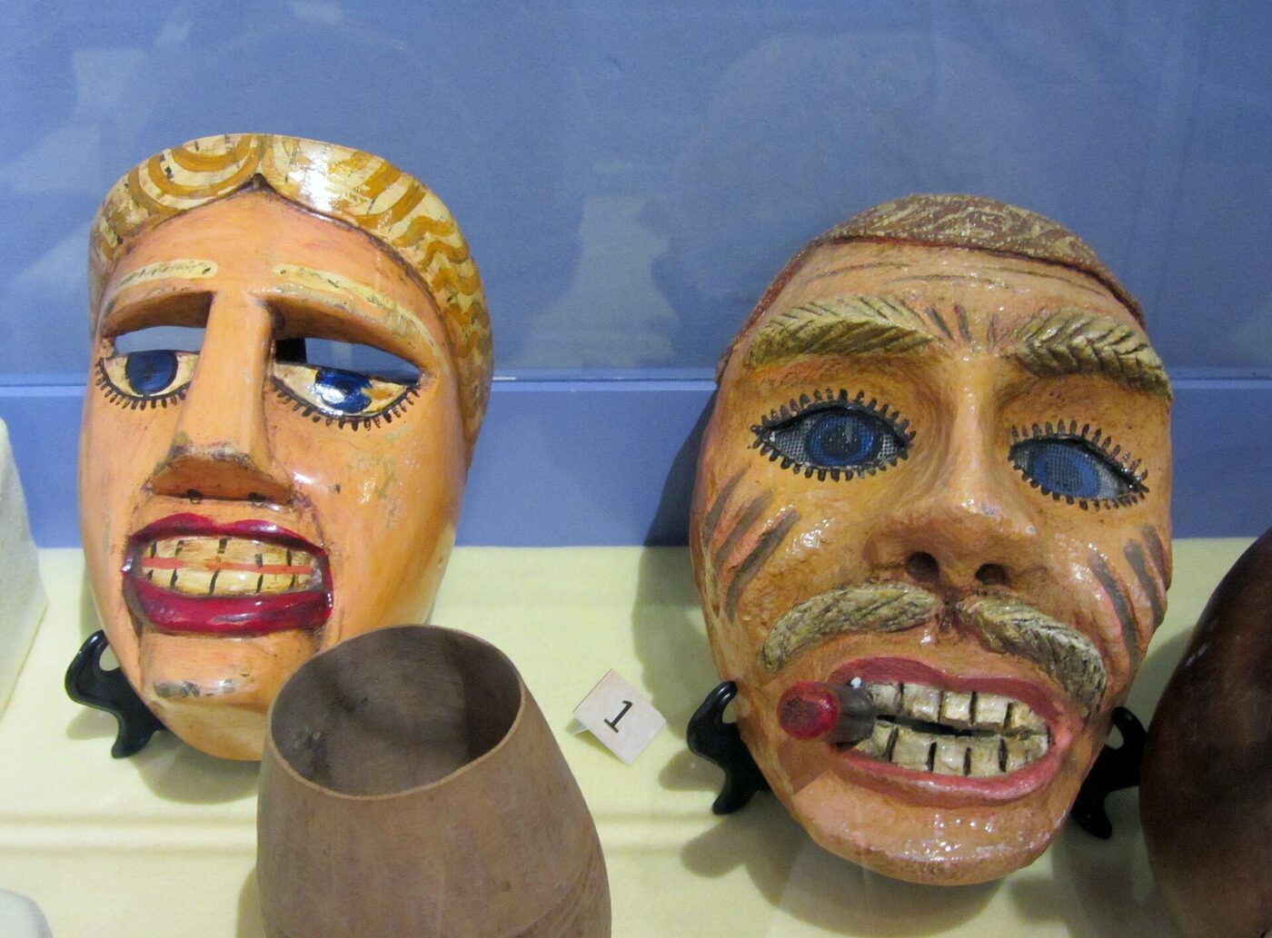 Two carved and painted wooden face masks on small stands in a museum case, one with red lips and blue eyes, the other with a grey moustache and a cigar gripped in its teeth, beside a plain gourd vessel and an exhibit label numbered one.
