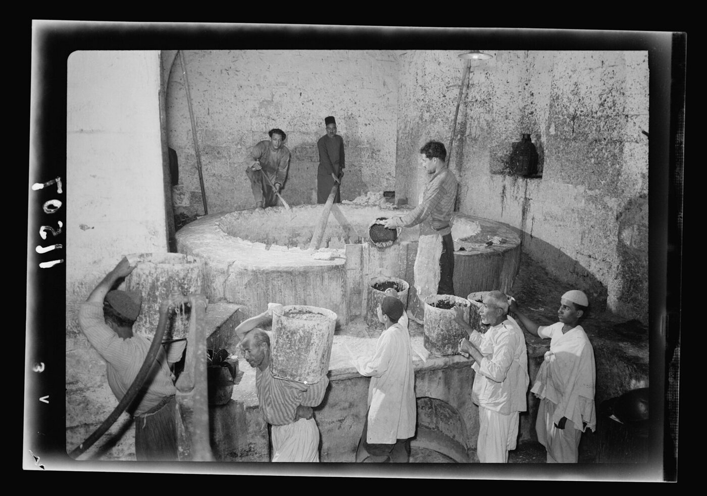 Black-and-white photograph of the interior of a stone-built soap works: a row of workers fill and shoulder buckets at the rim of a large circular boiling vat set into the floor, while two men stir it with long wooden paddles from the far side.