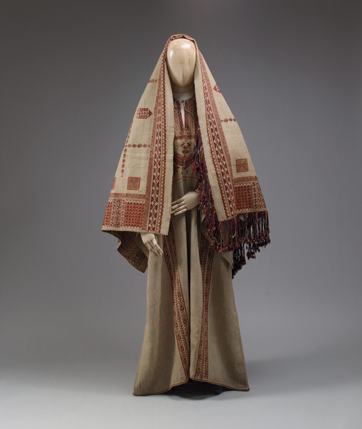 A long robe of natural-coloured linen on a mannequin, worn under a large head veil, both worked with dense red cross-stitch embroidery arranged in vertical bands and square panels, the veil ending in a knotted purple fringe.
