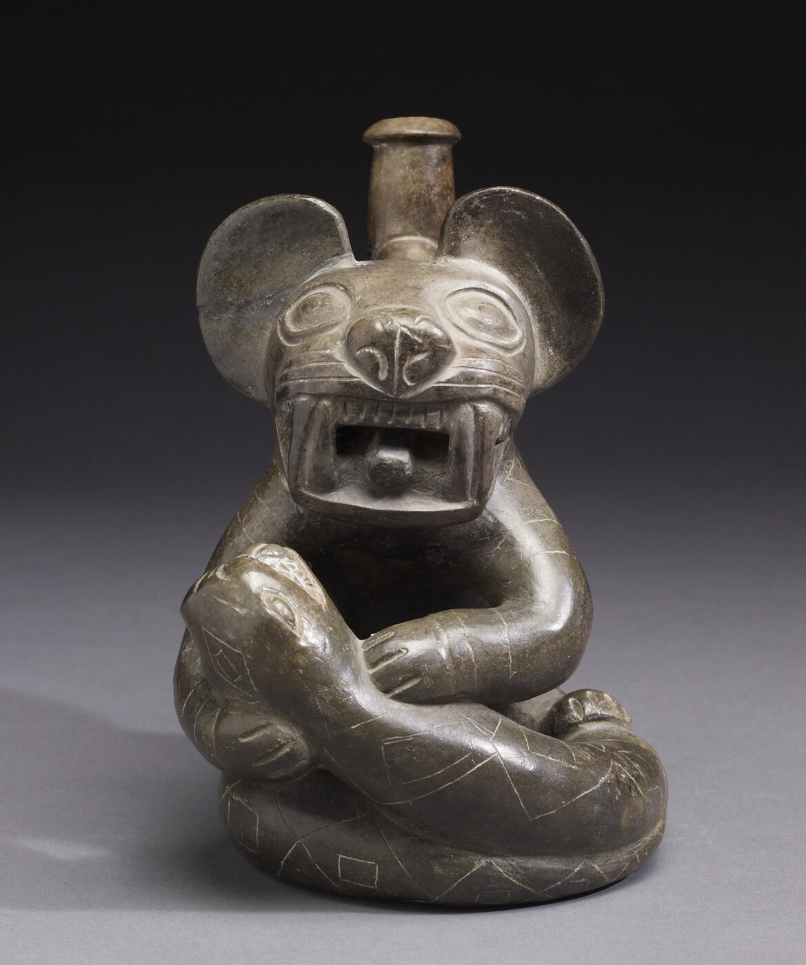 Dark grey-brown pre-Columbian ceramic vessel modelled as a seated cat-like animal with large round ears, bared teeth and an open mouth, its forelegs holding a small reptile-like creature, with a tubular stirrup spout rising behind the head, against a plain grey background.