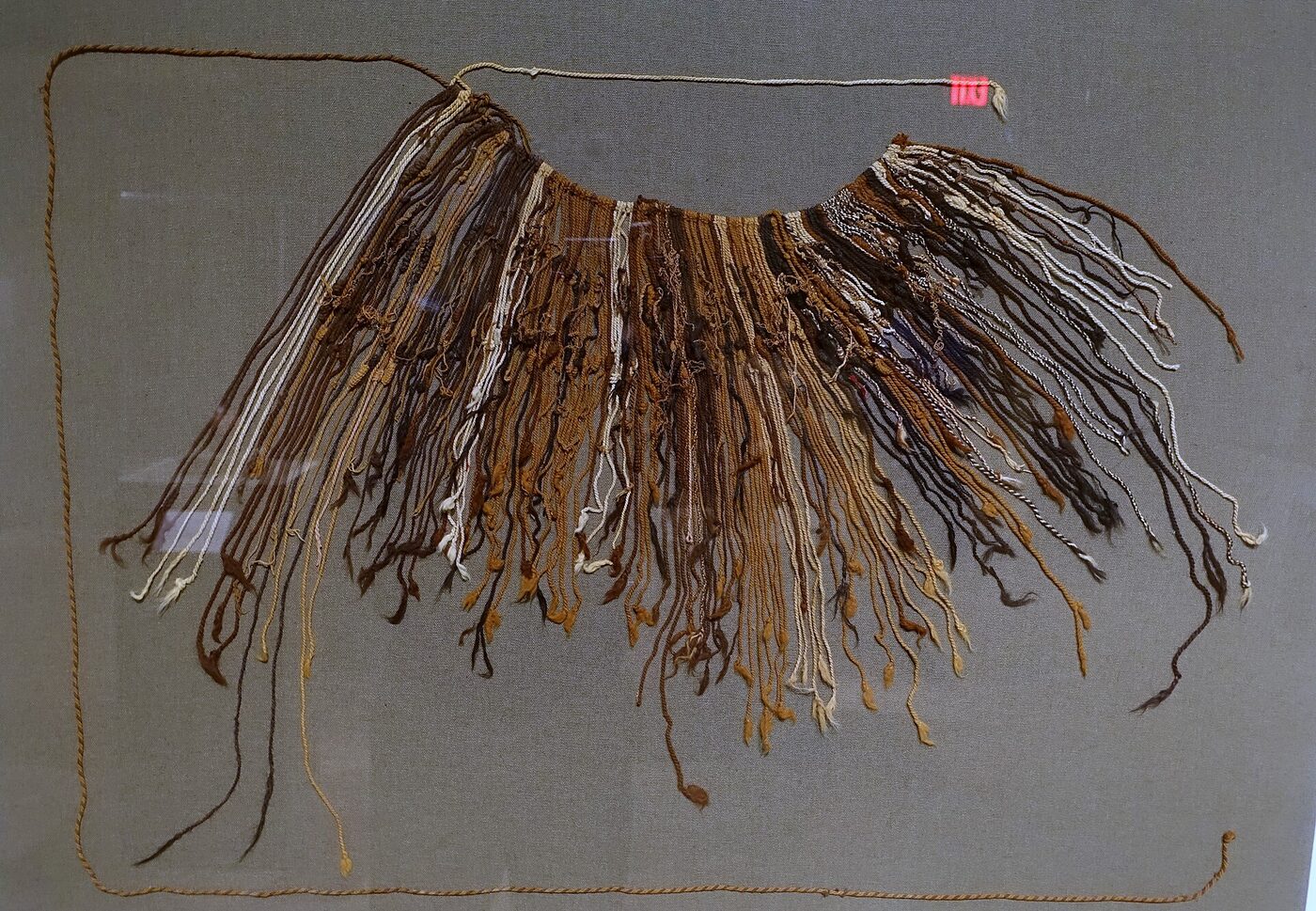 Dozens of faded brown, cream and dark cords hanging in a fan from a curved main cord, many with smaller subsidiary strings and knots tied along their length, mounted flat against grey fabric backing in a museum case.