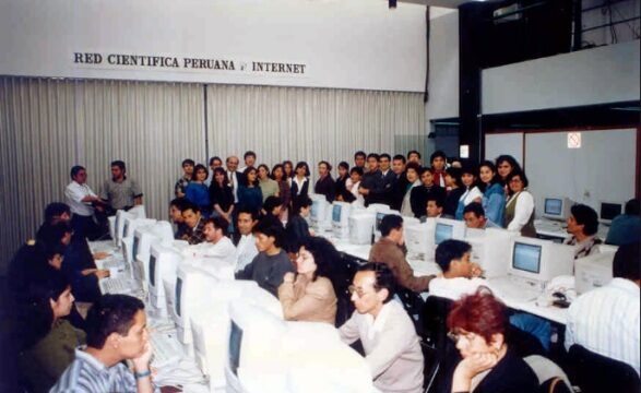 Black-and-white-toned colour photograph of a crowded room with rows of beige desktop computers on long tables, several dozen people seated at them and standing behind; a sign high on the wall reads RED CIENTIFICA PERUANA INTERNET.