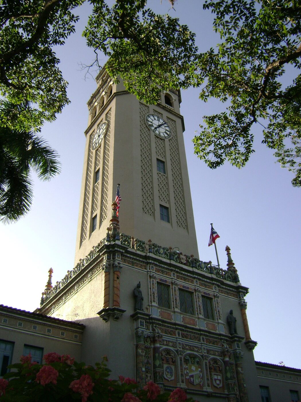 A tall pale clock tower with a latticed stone face, rising above an ornately tiled building front flanked by the United States and Puerto Rican flags, framed by tree branches.