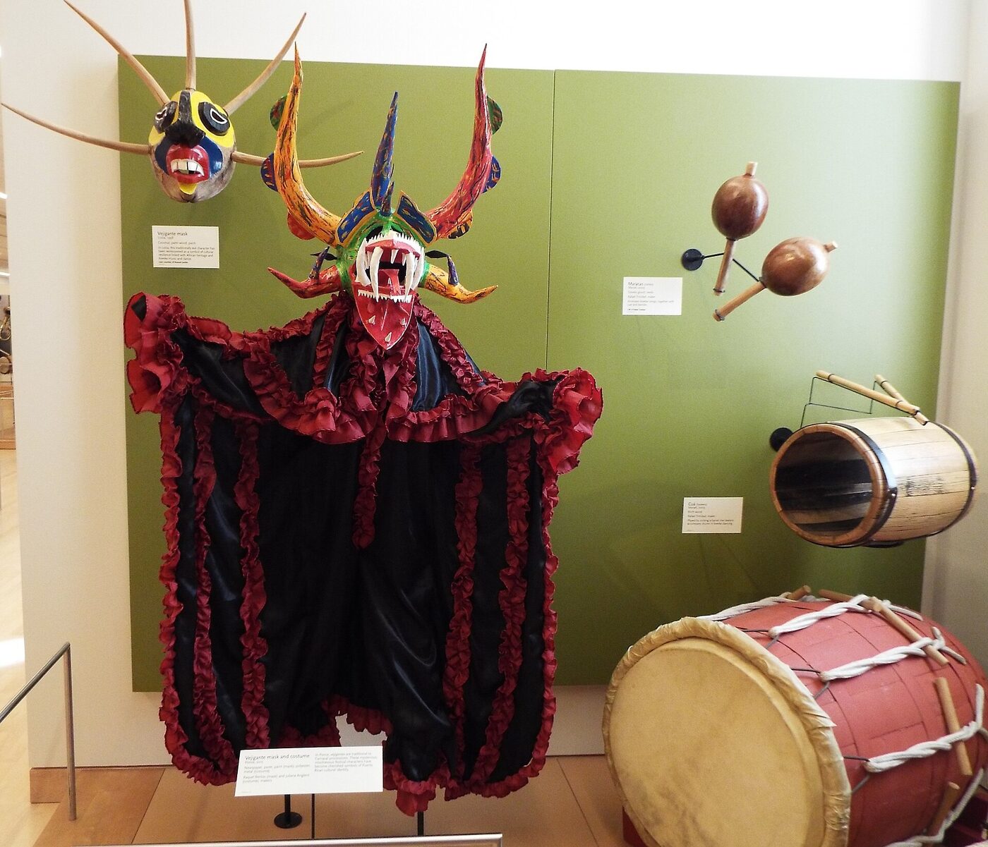 A brightly painted horned mask with bared teeth mounted above a long ruffled red and black costume in a museum gallery, with a smaller horned mask on the green wall behind and a barrel drum and maracas to the right.
