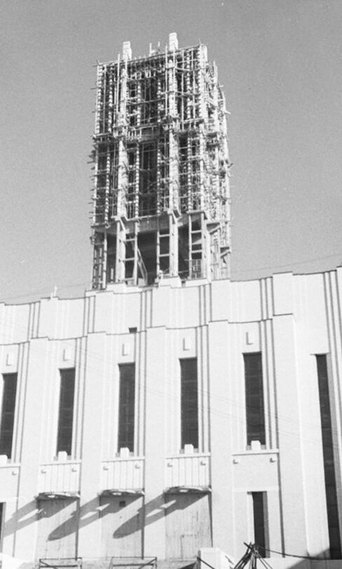 A black-and-white photograph looking up at a 1940s university building, the pale stone facade below and above it a tall tower still under scaffolding, its structural frame open against a plain sky.