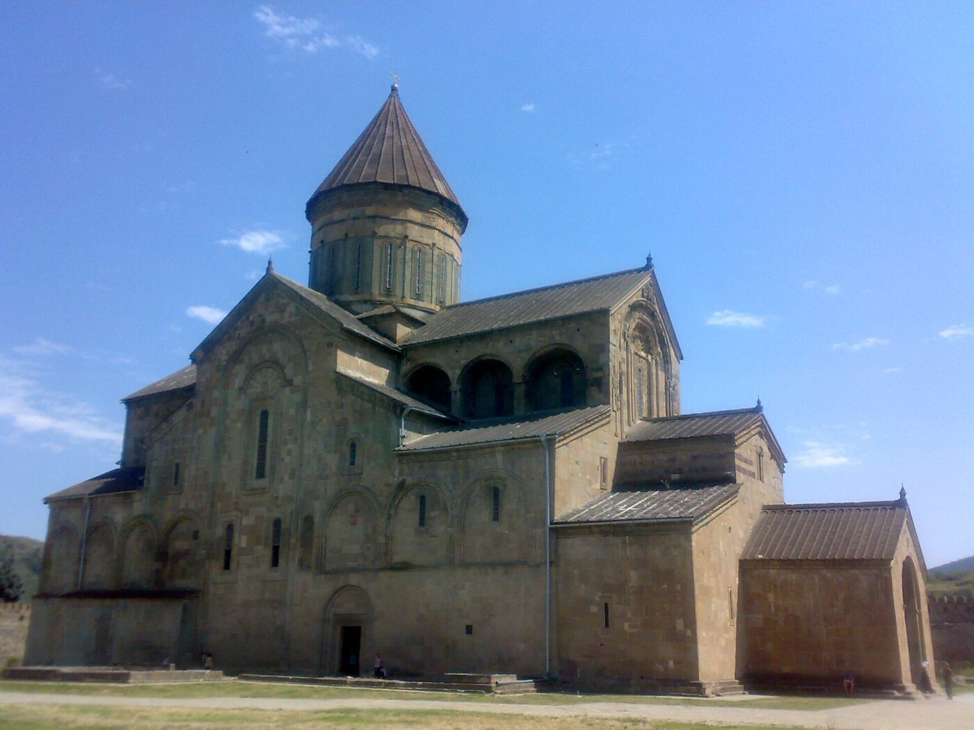 A large stone medieval Orthodox cathedral with a drum and conical dome, gabled roofs and blind arcading along its flank, seen from outside against a clear sky.