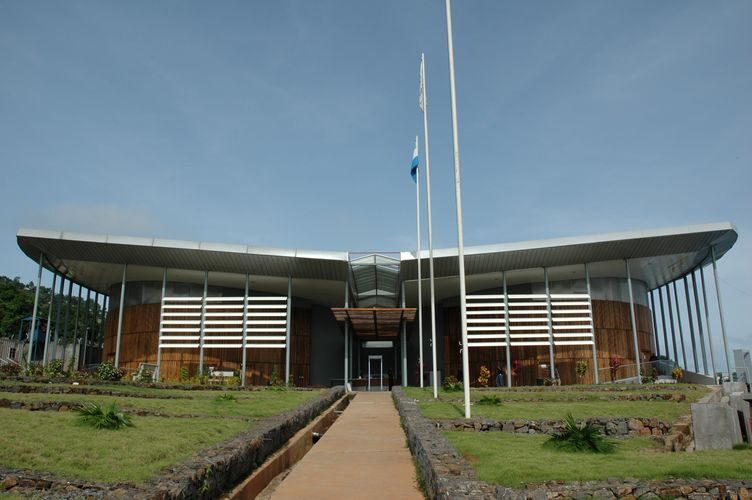 A low, wide modern building with a broad overhanging roof, timber-slatted end walls and a central entrance, approached by a paved path across a lawn, with three flagpoles in front of it, one flying a flag.