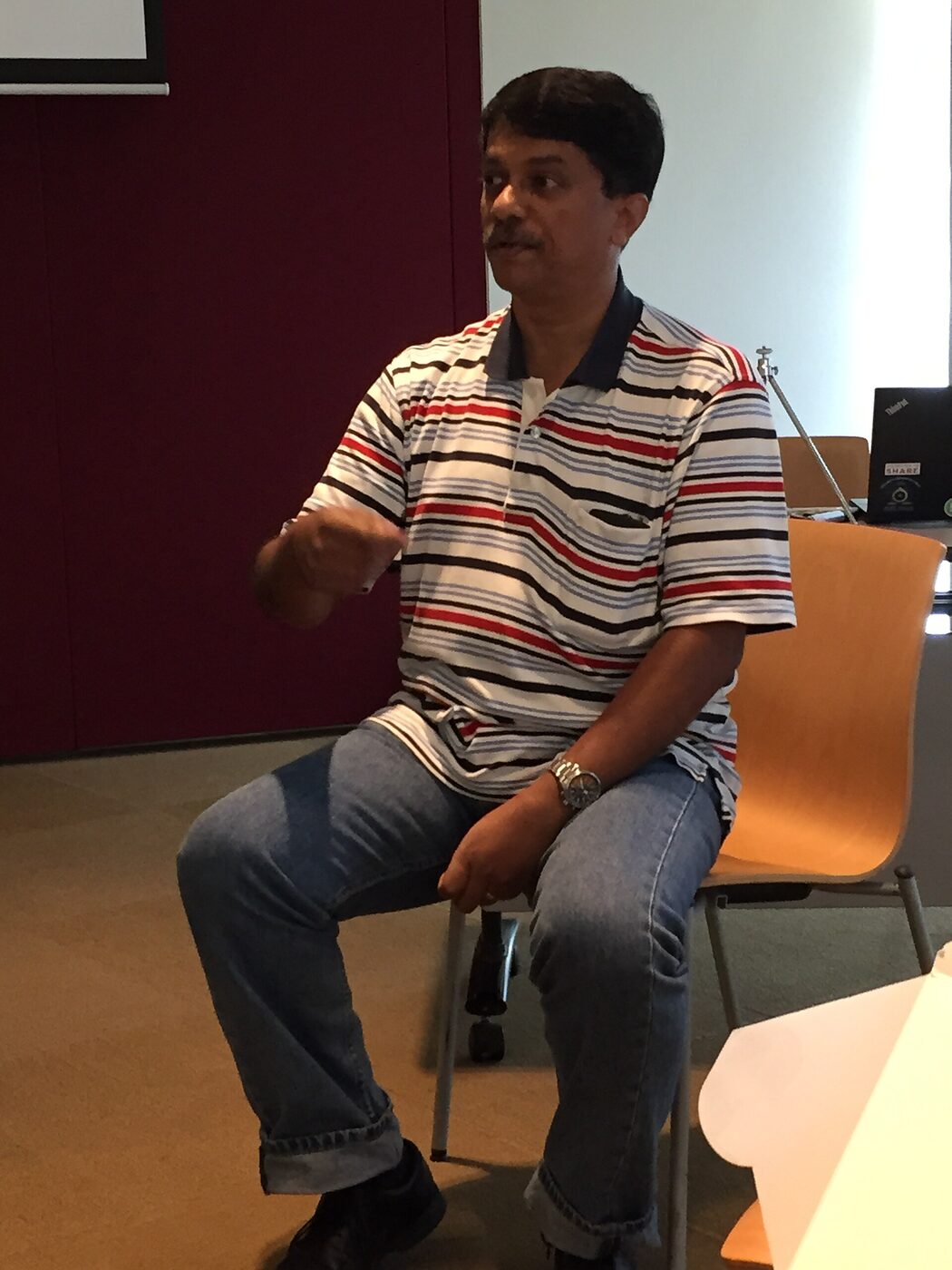 A seated man in a striped polo shirt gestures with one hand as he speaks, in a seminar room with a laptop on a table behind him.